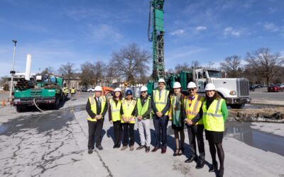 Construction works, both men and women, dressed in bright vests, stand at a worksite.
