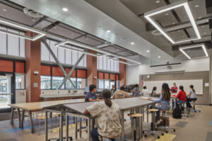 Students sit at large desks with a teacher presenting from the front of the room. Modern industrial lighting hangs from the ceiling, and large windows line the wall.